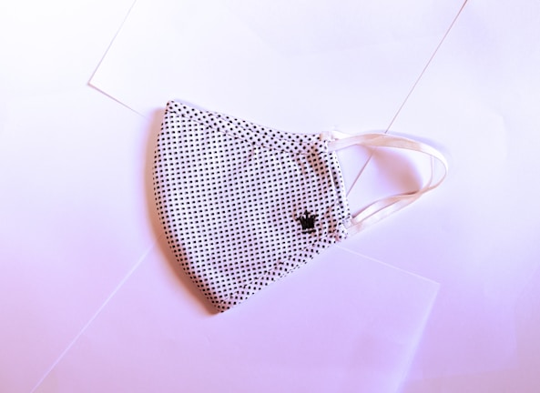 A fabric face mask with a black and white dotted pattern is laid flat on a slightly pink-tinted surface. The mask has elastic ear loops and a small embroidered crown detail on one side.