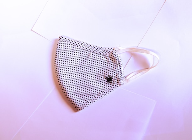 A fabric face mask with a black and white dotted pattern is laid flat on a slightly pink-tinted surface. The mask has elastic ear loops and a small embroidered crown detail on one side.
