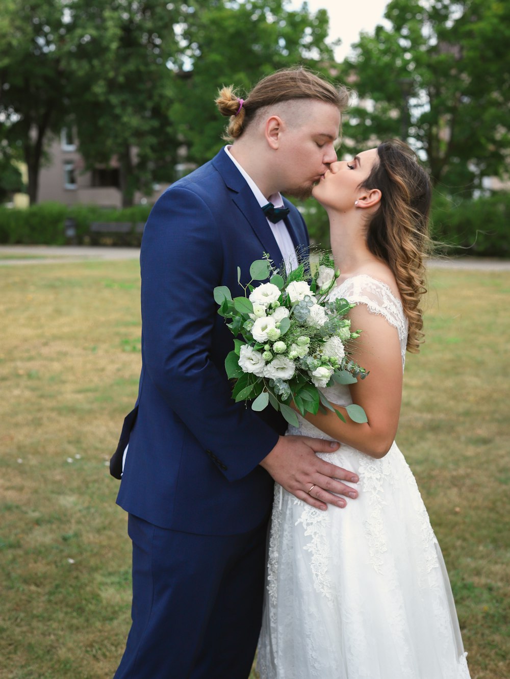 man in black suit kissing woman in white wedding dress on green grass field during daytime