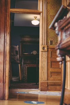 A well-lit, elegant interior with a view through a doorway into another room. The visible room features a classic wooden decor with ornate molding, and a partial view of a clock on a mantelpiece against a dark blue wall. A vintage lamp provides overhead lighting, casting a warm glow. The floor is wooden with a decorative rug, and the doorway features intricate wallpaper with a green floral pattern.
