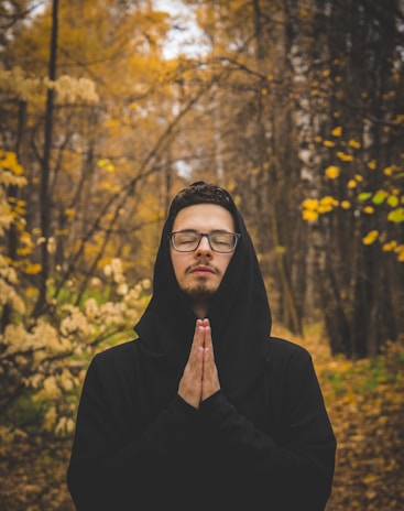 A person stands with closed eyes in a contemplative pose, hands together in prayer, in the middle of a forest during autumn. The trees around are covered with golden and brown leaves, creating a serene and peaceful environment.
