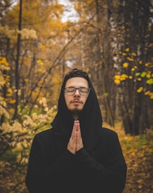 A person stands with closed eyes in a contemplative pose, hands together in prayer, in the middle of a forest during autumn. The trees around are covered with golden and brown leaves, creating a serene and peaceful environment.