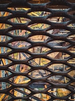 A complex arrangement of metal structures and grids is seen from an aerial perspective. The image displays intersecting beams and a lattice pattern that creates a geometric complexity. Sunlight casts shadows through the grid, contributing to the intricate visual pattern.