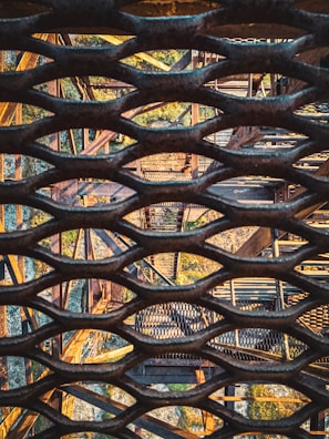 A complex arrangement of metal structures and grids is seen from an aerial perspective. The image displays intersecting beams and a lattice pattern that creates a geometric complexity. Sunlight casts shadows through the grid, contributing to the intricate visual pattern.