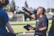 A man holding a book is speaking or teaching outdoors, gesturing with one hand while facing a group of people. They are in a grassy area with trees and a goal post in the background, suggesting a park or sports field setting.