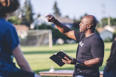 A man holding a book is speaking or teaching outdoors, gesturing with one hand while facing a group of people. They are in a grassy area with trees and a goal post in the background, suggesting a park or sports field setting.