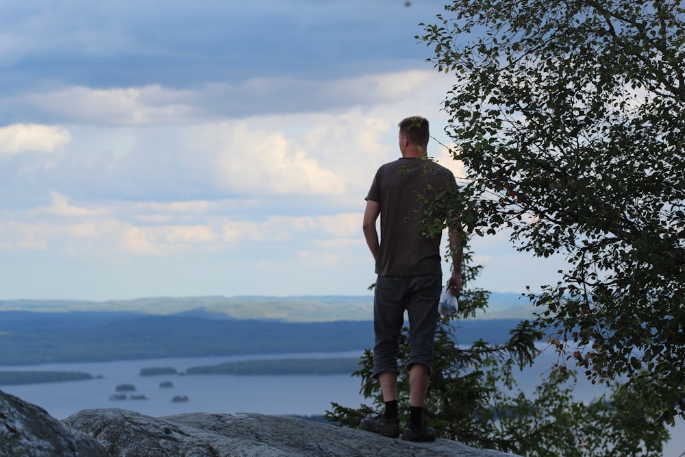 homme en t-shirt marron debout sur le rocher près de l’arbre vert pendant la journée