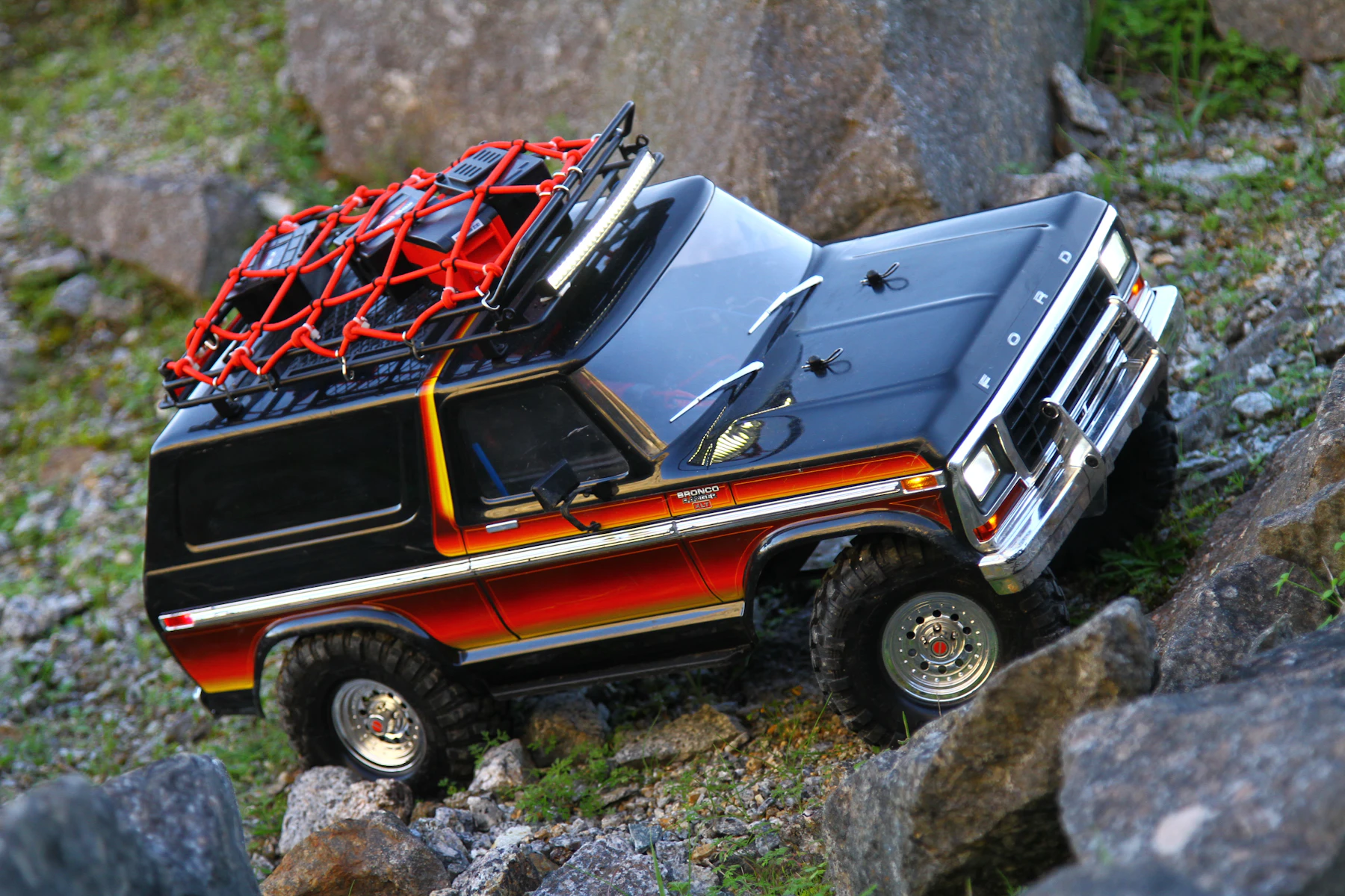 A black and orange scale trail truck with a loaded roof rack climbing between boulders
