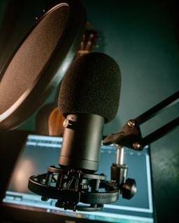 A close-up view of a black microphone with a pop filter mounted on an adjustable arm. The background features a blurred computer screen displaying editing software, suggesting a recording or audio editing setup.
