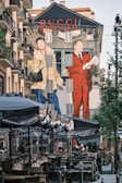 A large mural on the side of a building features two fashionable figures, one in a fur coat and the other in a red suit, promoting the Gucci brand. The scene is set in an urban environment with outdoor seating under umbrellas and people walking by. The wall art is vibrant and eye-catching, contributing to a lively street atmosphere.