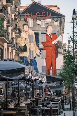 A large mural on the side of a building features two fashionable figures, one in a fur coat and the other in a red suit, promoting the Gucci brand. The scene is set in an urban environment with outdoor seating under umbrellas and people walking by. The wall art is vibrant and eye-catching, contributing to a lively street atmosphere.