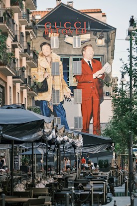 A large mural on the side of a building features two fashionable figures, one in a fur coat and the other in a red suit, promoting the Gucci brand. The scene is set in an urban environment with outdoor seating under umbrellas and people walking by. The wall art is vibrant and eye-catching, contributing to a lively street atmosphere.