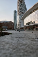 A modern urban landscape with distinctive architectural structures. Prominent buildings feature a cylindrical tower with a lattice design, a curved facade with reflective glass windows, and a wooden dome-shaped structure. The perspective is ground-level, highlighting the walkway with concrete tiles leading towards the buildings.