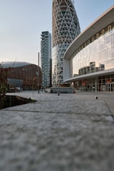 A modern urban landscape with distinctive architectural structures. Prominent buildings feature a cylindrical tower with a lattice design, a curved facade with reflective glass windows, and a wooden dome-shaped structure. The perspective is ground-level, highlighting the walkway with concrete tiles leading towards the buildings.