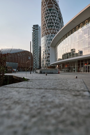 A modern urban landscape with distinctive architectural structures. Prominent buildings feature a cylindrical tower with a lattice design, a curved facade with reflective glass windows, and a wooden dome-shaped structure. The perspective is ground-level, highlighting the walkway with concrete tiles leading towards the buildings.