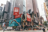 A busy urban scene with towering skyscrapers and numerous large advertisements. The billboards feature health-related messages and images of people engaged in athletic activities. Vehicles and pedestrians move along the street below, with iconic city taxicabs visible.