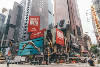 A busy urban scene with towering skyscrapers and numerous large advertisements. The billboards feature health-related messages and images of people engaged in athletic activities. Vehicles and pedestrians move along the street below, with iconic city taxicabs visible.