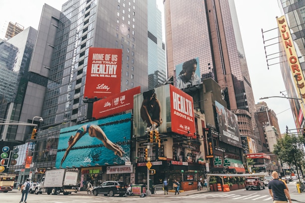 A busy urban scene with towering skyscrapers and numerous large advertisements. The billboards feature health-related messages and images of people engaged in athletic activities. Vehicles and pedestrians move along the street below, with iconic city taxicabs visible.
