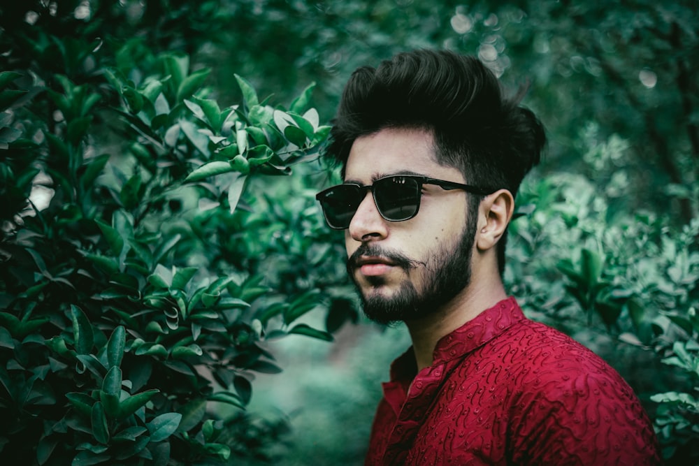 man in red crew neck shirt wearing black sunglasses standing beside green leaves during daytime