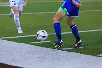 Two soccer players in action, focusing on their legs and feet as they compete for control of the ball on a grass field. One player in a blue kit is about to kick the ball while the other in white approaches.