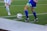 Two soccer players in action, focusing on their legs and feet as they compete for control of the ball on a grass field. One player in a blue kit is about to kick the ball while the other in white approaches.
