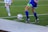 Two soccer players in action, focusing on their legs and feet as they compete for control of the ball on a grass field. One player in a blue kit is about to kick the ball while the other in white approaches.