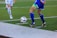 Two soccer players in action, focusing on their legs and feet as they compete for control of the ball on a grass field. One player in a blue kit is about to kick the ball while the other in white approaches.