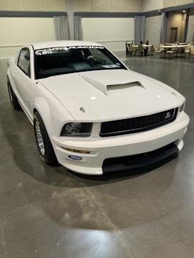 A white Ford Mustang with racing decals is parked indoors on a polished concrete floor. The car features a hood scoop and various stickers, including Ford Racing branding. In the background, there are several tables and chairs arranged along the walls, suggesting a convention or exhibition setting.