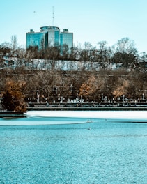 A large, modern building with glass windows stands on a hill surrounded by trees. The trees appear bare, suggesting winter or early spring. A calm body of water stretches across the foreground, partially frozen, with birds resting on the ice. The letters 'TABRIZ' are visible on the hillside, indicating the location.