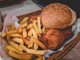 A woven basket contains a meal consisting of crispy golden French fries, a piece of fried chicken, and a sesame seed hamburger bun.