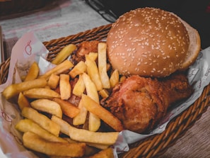 A woven basket contains a meal consisting of crispy golden French fries, a piece of fried chicken, and a sesame seed hamburger bun.