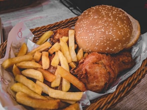 A woven basket contains a meal consisting of crispy golden French fries, a piece of fried chicken, and a sesame seed hamburger bun.