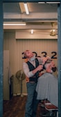 A barber wearing a mask is giving a haircut to a client who is also masked. The setting is a well-lit, modern barber shop with wooden flooring. The barber is using scissors to cut the client's hair, and the client is seated in a chair covered with a striped cape. In the background, there is a wall with numbers and a vintage-style fan.