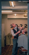 A barber wearing a mask is giving a haircut to a client who is also masked. The setting is a well-lit, modern barber shop with wooden flooring. The barber is using scissors to cut the client's hair, and the client is seated in a chair covered with a striped cape. In the background, there is a wall with numbers and a vintage-style fan.