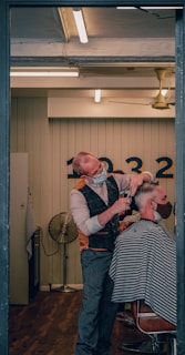 A barber wearing a mask is giving a haircut to a client who is also masked. The setting is a well-lit, modern barber shop with wooden flooring. The barber is using scissors to cut the client's hair, and the client is seated in a chair covered with a striped cape. In the background, there is a wall with numbers and a vintage-style fan.