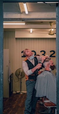 A barber wearing a mask is giving a haircut to a client who is also masked. The setting is a well-lit, modern barber shop with wooden flooring. The barber is using scissors to cut the client's hair, and the client is seated in a chair covered with a striped cape. In the background, there is a wall with numbers and a vintage-style fan.