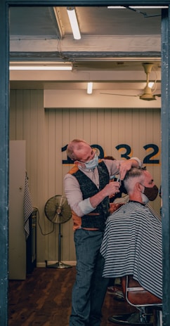 A barber wearing a mask is giving a haircut to a client who is also masked. The setting is a well-lit, modern barber shop with wooden flooring. The barber is using scissors to cut the client's hair, and the client is seated in a chair covered with a striped cape. In the background, there is a wall with numbers and a vintage-style fan.