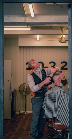 A barber wearing a mask is giving a haircut to a client who is also masked. The setting is a well-lit, modern barber shop with wooden flooring. The barber is using scissors to cut the client's hair, and the client is seated in a chair covered with a striped cape. In the background, there is a wall with numbers and a vintage-style fan.