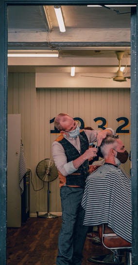 A barber wearing a mask is giving a haircut to a client who is also masked. The setting is a well-lit, modern barber shop with wooden flooring. The barber is using scissors to cut the client's hair, and the client is seated in a chair covered with a striped cape. In the background, there is a wall with numbers and a vintage-style fan.