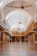 An ornate and grand library with high arched ceilings and intricate architectural details. The walls are lined with tall bookshelves filled with numerous books, and the floor features a polished, patterned design. Chandeliers hang from the ceiling, providing a sense of elegance and sophistication.