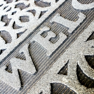 A close-up of a welcome mat with textured, raised lettering on a rough surface. The mat features decorative patterns and appears worn, indicating frequent use.