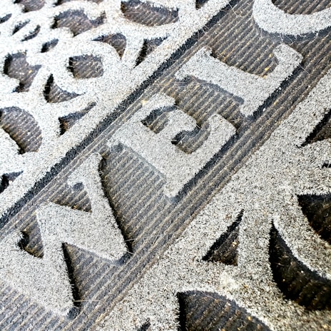 A close-up of a welcome mat with textured, raised lettering on a rough surface. The mat features decorative patterns and appears worn, indicating frequent use.