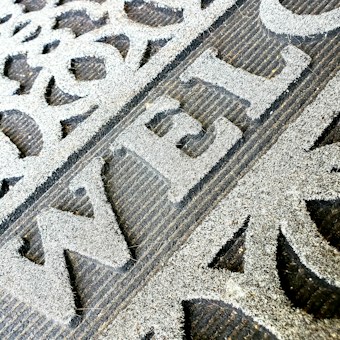 A close-up of a welcome mat with textured, raised lettering on a rough surface. The mat features decorative patterns and appears worn, indicating frequent use.