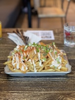 A plate of loaded French fries topped with melted cheese, sour cream, chopped green onions, and sprinkled with herbs. The dish is placed on a rustic wooden table next to a napkin and cutlery, with a glass of water nearby.