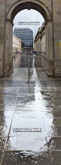 An arched stone gateway leads to a street flanked by buildings on both sides. Above the arch, the words 'MERCHANT CITY' are displayed. The ground is wet, reflecting the sign and creating a mirror effect. Towards the background, a modern glass building stands amidst older structures.