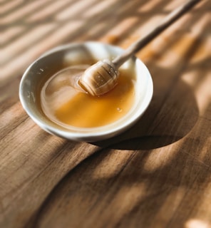 A bowl filled with golden honey, accompanied by a wooden honey dipper resting inside. The bowl sits on a wooden surface with soft, warm lighting casting gentle shadows.