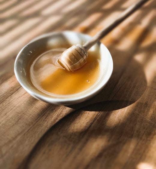 A bowl filled with golden honey, accompanied by a wooden honey dipper resting inside. The bowl sits on a wooden surface with soft, warm lighting casting gentle shadows.