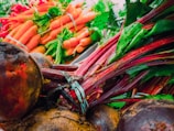 A vibrant pile of fresh vegetables including beets with leafy green tops and bright orange carrots with green stems. The produce looks freshly harvested and is arranged in a way that highlights their natural colors and textures. The leafy greens and rich orange hues create a dynamic and colorful composition.