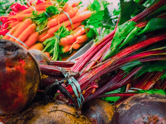 A vibrant pile of fresh vegetables including beets with leafy green tops and bright orange carrots with green stems. The produce looks freshly harvested and is arranged in a way that highlights their natural colors and textures. The leafy greens and rich orange hues create a dynamic and colorful composition.