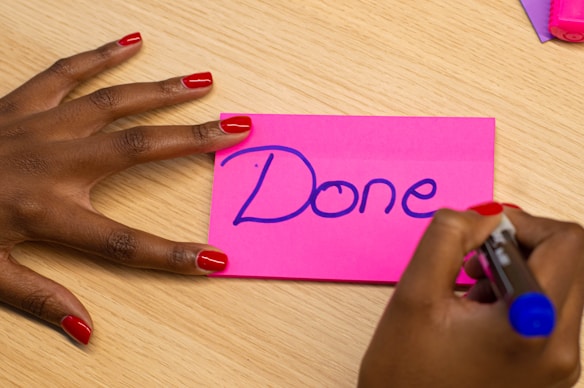 A hand with red-painted nails holds a pink sticky note on a wooden surface. The word 'Done' is written in blue marker on the note. Another hand with similar nail polish is holding a blue marker, finishing writing or drawing.